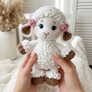 May include: A handmade white crochet lamb doll with brown hooves and ear details. The lamb has pink accents on its ears and cheeks, and black eyes. The doll is held by a person.