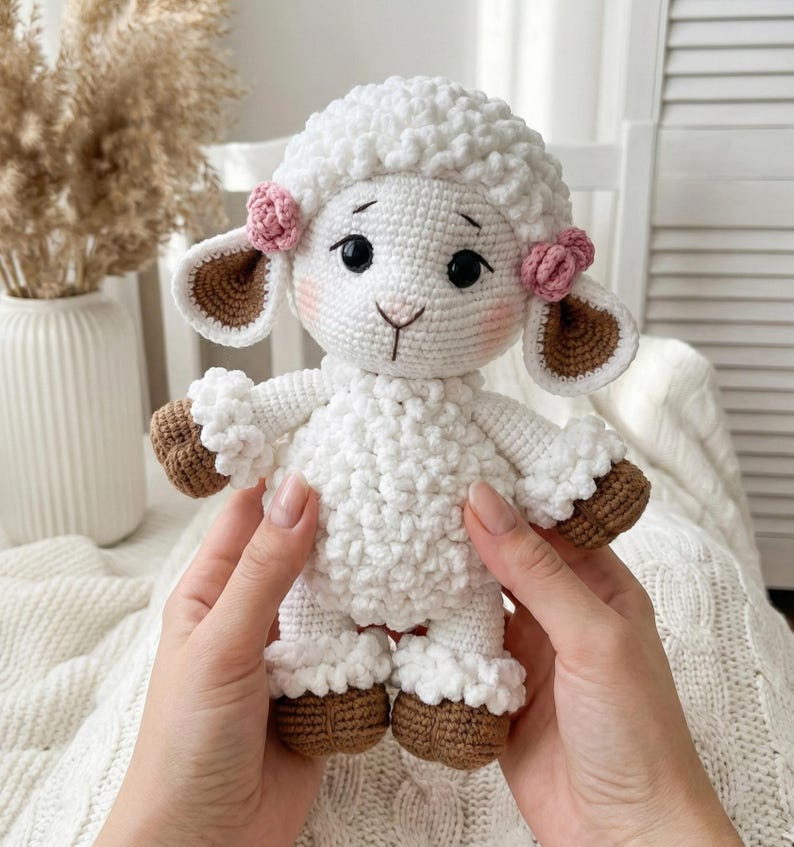 1) Crochet lamb doll with curly white fleece and brown hooves held in two hands
2) Front view of amigurumi sheep showing floppy ears, safety eyes, and stitched smile detail
3) Close-up of crochet lamb face with pink cheeks, small nose, and flower accents by the ears
4) Detail of lamb body showing textured bobble fleece and ruffled cuffs on arms and legs
5) Handmade crochet sheep plush photographed in a cozy neutral nursery setting with soft blanket background
