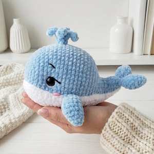 May include: A light blue crocheted whale plush toy with a white belly, black eyes, and a pink blush. The whale has a water spout on top and flipper-like fins. The plush is held in a person's hand.