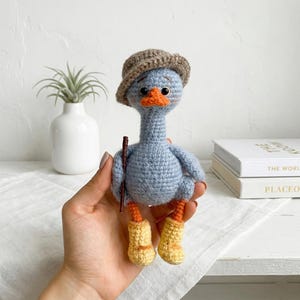 May include: A handmade crochet goose doll in light blue, wearing a brown hat and yellow boots. The goose has an orange beak and feet, and holds a small brown stick. The doll is a whimsical, handcrafted item.
