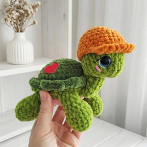 Puede incluir: Una tortuga verde hecha a mano con ganchillo, con un corazón rojo en la espalda y un sombrero naranja. La tortuga tiene grandes ojos azules y mejillas rosadas. El peluche se sostiene en una mano.