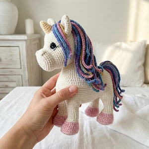 May include: A cream-coloured crocheted unicorn toy with a tan horn and pink hooves. The unicorn features a flowing mane and tail in shades of blue, purple, and yellow. The toy is handmade with black eyes.