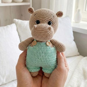 ALT TEXT (5)
1) Crochet hippo doll with beige yarn body and black safety eyes, wearing green overalls with two wooden-style buttons, held in hands.
2) Close-up of amigurumi hippo face showing rounded snout shaping, tiny ears, and glossy safety eyes.
3) Front view of crochet hippo plush in green overalls, showing strap details and button accents on the chest.
4) Nursery-style photo of the crocheted hippo toy held in hands with cozy neutral decor, wooden toys, and dried florals in the background.
