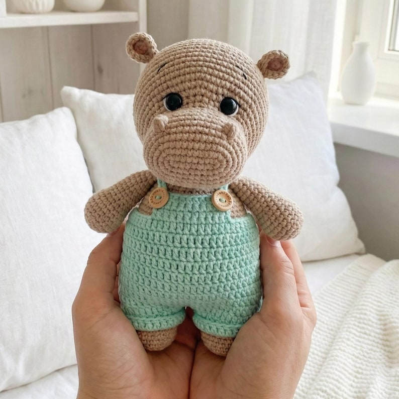 ALT TEXT (5)
1) Crochet hippo doll with beige yarn body and black safety eyes, wearing green overalls with two wooden-style buttons, held in hands.
2) Close-up of amigurumi hippo face showing rounded snout shaping, tiny ears, and glossy safety eyes.
3) Front view of crochet hippo plush in green overalls, showing strap details and button accents on the chest.
4) Nursery-style photo of the crocheted hippo toy held in hands with cozy neutral decor, wooden toys, and dried florals in the background.