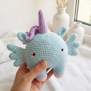 Puede incluir: Un juguete de unicornio de ganchillo hecho a mano en azul claro, lavanda y rosa. El unicornio tiene alas, un cuerno y pequeños ojos negros. El juguete mide aproximadamente 15 cm de alto.