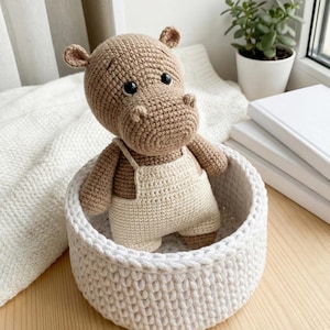 May include: A crocheted hippo toy in a white basket. The hippo is light brown with black eyes and wears cream-colored overalls. The basket is round and has a textured, woven appearance. A plant in a white pot and white books are in the background.