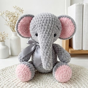 Könnte beinhalten: Ein handgefertigtes, graues Häkelelefant-Plüschtier mit rosa Akzenten an Ohren und Füßen. Der Elefant hat ein graues Band, das zu einer Schleife um den Hals gebunden ist, und schwarze Knopfaugen. Das Spielzeug sitzt auf einer weißen, strukturierten Oberfläche.