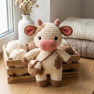 May include: A cream and brown crochet cow plush toy with pink accents on the nose and ears. The cow has a small brown purse. It is positioned in front of a wooden crate filled with balls of yarn.