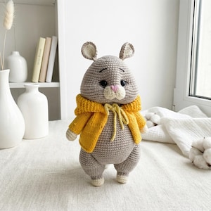 May include: A handmade grey crochet bunny wearing a yellow jacket. The bunny has large black eyes, a pink nose, and small, upright ears. The jacket is a bright yellow with a matching tie closure. The bunny is standing on a white surface.