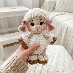 1) Crochet lamb doll with curly white fleece and brown hooves held in two hands
2) Front view of amigurumi sheep showing floppy ears, safety eyes, and stitched smile detail
3) Close-up of crochet lamb face with pink cheeks, small nose, and flower accents by the ears
4) Detail of lamb body showing textured bobble fleece and ruffled cuffs on arms and legs
5) Handmade crochet sheep plush photographed in a cozy neutral nursery setting with soft blanket background