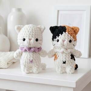 May include: Two handmade crochet cat dolls. One is white with a lavender collar, the other is calico with black, orange, and white patches. Both have black eyes and pink noses. They are standing on a white surface.