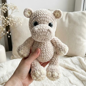 May include: A handmade, light beige crocheted hippo plush toy. The hippo has large, dark eyes and small, rounded ears. Its body is soft and textured, with lighter beige accents on the feet and ears. The plush is held in a hand.