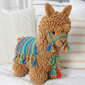 May include: A handmade brown crochet llama with a colorful blanket and tassels. The llama has a blue halter and colorful tassels. The llama is standing on a white surface with pillows in the background.