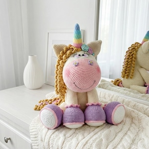 May include: A handmade crochet unicorn doll with a rainbow horn, flowing golden mane, and pink cheeks. The unicorn has purple and white hooves and is sitting on a white surface. The doll is a whimsical and charming decorative item.