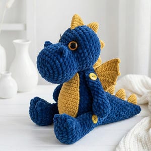 Puede incluir: Un peluche de dragón de ganchillo azul y amarillo hecho a mano. El dragón tiene ojos grandes y redondos, alas amarillas y detalles amarillos en la cabeza y la cola. Está sobre una superficie blanca.