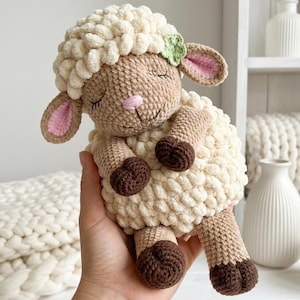 Puede incluir: Un juguete de oveja de ganchillo hecho a mano en crema, marrón y beige. La oveja tiene un cuerpo esponjoso, una cara beige y orejas forradas de rosa. Una pequeña flor verde adorna su cabeza. El juguete es suave y tierno.