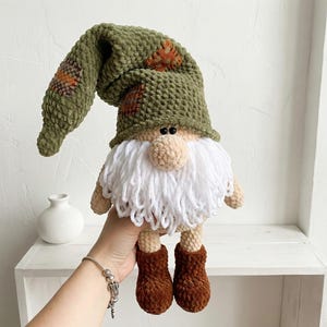 Puede incluir: Una muñeca de gnomo de peluche hecha a mano con un sombrero largo verde oliva adornado con parches naranjas y marrones. El gnomo tiene una barba blanca, una nariz beige y botas marrones. La muñeca mide aproximadamente 25 cm de alto.