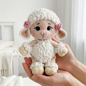 1) Crochet lamb doll with curly white fleece and brown hooves held in two hands
2) Front view of amigurumi sheep showing floppy ears, safety eyes, and stitched smile detail
3) Close-up of crochet lamb face with pink cheeks, small nose, and flower accents by the ears
4) Detail of lamb body showing textured bobble fleece and ruffled cuffs on arms and legs
5) Handmade crochet sheep plush photographed in a cozy neutral nursery setting with soft blanket background