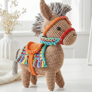 Op de afbeelding: Een gehaakte ezelbeeldje in bruin-, grijs-, oranje-, rood- en blauwtinten. De ezel heeft een zadel, een halster en een manen. Het zadel is oranje en de deken is veelkleurig met kwastjes. De ezel staat op een wit oppervlak.