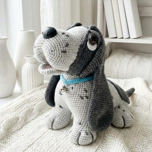 Może przedstawiać: Ręcznie robiony szydełkowy pies w odcieniach szarości i bieli, z długimi, oklapniętymi uszami i niebieską obrożą z małym dzwoneczkiem. Pies siedzi na białym, teksturowanym kocu. Pies ma duże brązowe oczy.