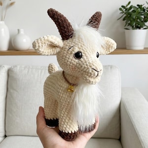 May include: A handmade crochet goat with a cream-colored body, brown horns, and black eyes. It has a white fluffy beard and a small gold bell around its neck. The goat is a soft sculpture, perfect for nursery decor or a child's toy.