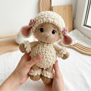1) Crochet lamb doll with curly white fleece and brown hooves held in two hands
2) Front view of amigurumi sheep showing floppy ears, safety eyes, and stitched smile detail
3) Close-up of crochet lamb face with pink cheeks, small nose, and flower accents by the ears
4) Detail of lamb body showing textured bobble fleece and ruffled cuffs on arms and legs
5) Handmade crochet sheep plush photographed in a cozy neutral nursery setting with soft blanket background