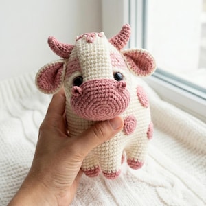 Puede incluir: Un peluche de vaca de ganchillo hecho a mano en crema y rosa. La vaca tiene manchas, orejas, cuernos y hocico rosas. El peluche se sostiene en una mano sobre un fondo blanco texturizado.