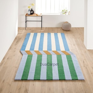 May include: A rectangular rug with a striped design in shades of blue, white, and green. The rug features a unique geometric pattern and the text "DuaCarpet". The rug is placed on a wooden floor.