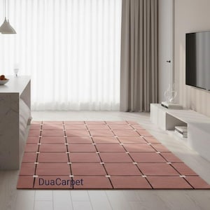 Könnte beinhalten: Ein rechteckiger, rosafarbener Teppich mit einem Rastermuster aus Quadraten und dünnen schwarzen Linien. Der Teppich befindet sich in einem modernen Wohnzimmer mit einer weißen Arbeitsplatte, Vorhängen und einem Fernseher. Der Text "DuaCarpet" ist sichtbar.