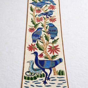 May include: Embroidered textile runner with a tree design, featuring blue birds, red flowers, and green leaves. The birds are of different sizes and poses. The runner has a cream background, brown border, and white fringe.