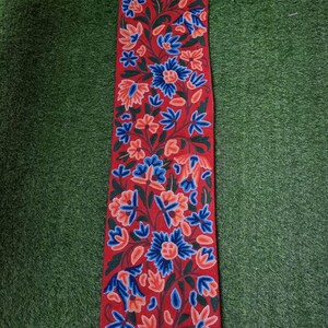 Puede incluir: Un textil rectangular rojo con un estampado floral. El diseño presenta flores azules y naranjas con hojas y tallos verdes. Flecos blancos adornan el borde inferior y borlas blancas en la parte superior. Probablemente sea una bufanda o un camino de mesa.