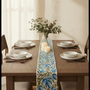 Puede incluir: Una mesa de comedor de madera para cuatro personas con platos, cuencos y cubiertos blancos. Un camino de mesa floral azul y verde con borlas recorre el centro. Un jarrón con vegetación y velas encendidas decoran la mesa.