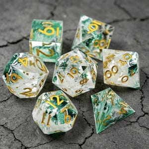 Könnte beinhalten: Set aus sieben polyedrischen Würfeln für Tabletop-Spiele. Die Würfel sind transparent mit einem grün-weißen Marmoreffekt und goldenen Zahlen. Enthält einen D4, D6, D8, D10, D12, D20 und einen Prozentwürfel.