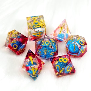May include: A set of seven polyhedral dice for tabletop games. Each die is clear with blue and red accents, gold glitter, and gold numbering. The dice are arranged on a white, fluffy surface.