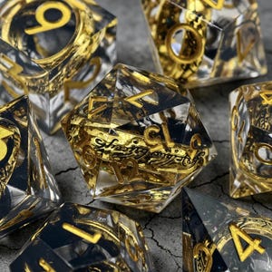 May include: A set of clear, multi-sided dice featuring gold numerals and intricate gold detailing within. The dice are displayed on a grey surface, with some in sharp focus and others slightly blurred.