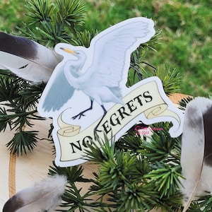 May include: A white egret with its wings spread wide, perched on a scroll with the text "No Regrets". The sticker is on a green pine branch.
