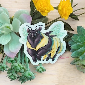May include: A sticker featuring a black bear with yellow and white stripes, resembling a bumblebee, with translucent green wings. The sticker is on a light blue background.