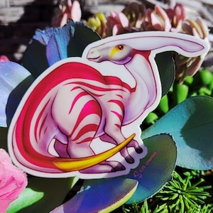 May include: A pink and white striped dinosaur sticker with a yellow tail and a white head. The sticker is on a green and brown background.