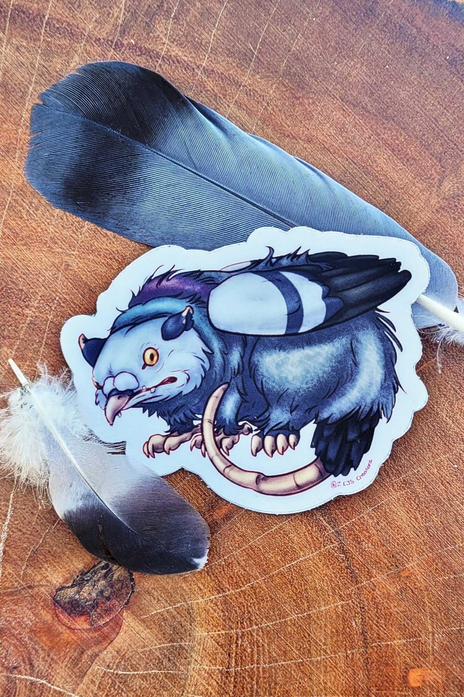 A sticker, staged with feathers, featuring a gryphon that is a hybrid of a pigeon and a possum.
