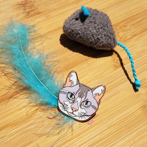 May include: A grey cat head enamel pin with blue eyes and a white stripe on its forehead. The pin is on a wooden surface with a blue feather and a grey plush toy mouse with a blue tail.