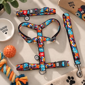 Könnte beinhalten: Ein Haustiergeschirr-, Halsband- und Leinenset mit einem bunten Monstermuster in Blau-, Orange- und Gelbtönen. Das Set enthält schwarze Kunststoffschnallen, Metallringe und kleine runde Anhänger mit QR-Codes. Der Markenname WAU DOG ist auf roten Stoffetiketten am Geschirr und Halsband aufgedruckt.