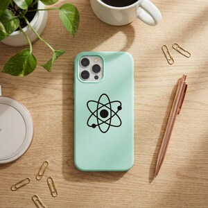 May include: A pale green phone case featuring a black atom symbol. A rose gold pen, gold paper clips, a white coffee cup, a plant, and a wireless charger are on a wooden surface.