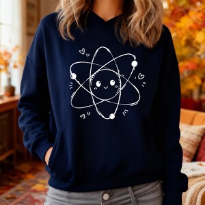 May include: A navy blue hooded sweatshirt with a white atom graphic. The atom design has a smiling face at the centre, with orbiting circles and dots, and small heart details. The sweatshirt has a front pocket and a hood.