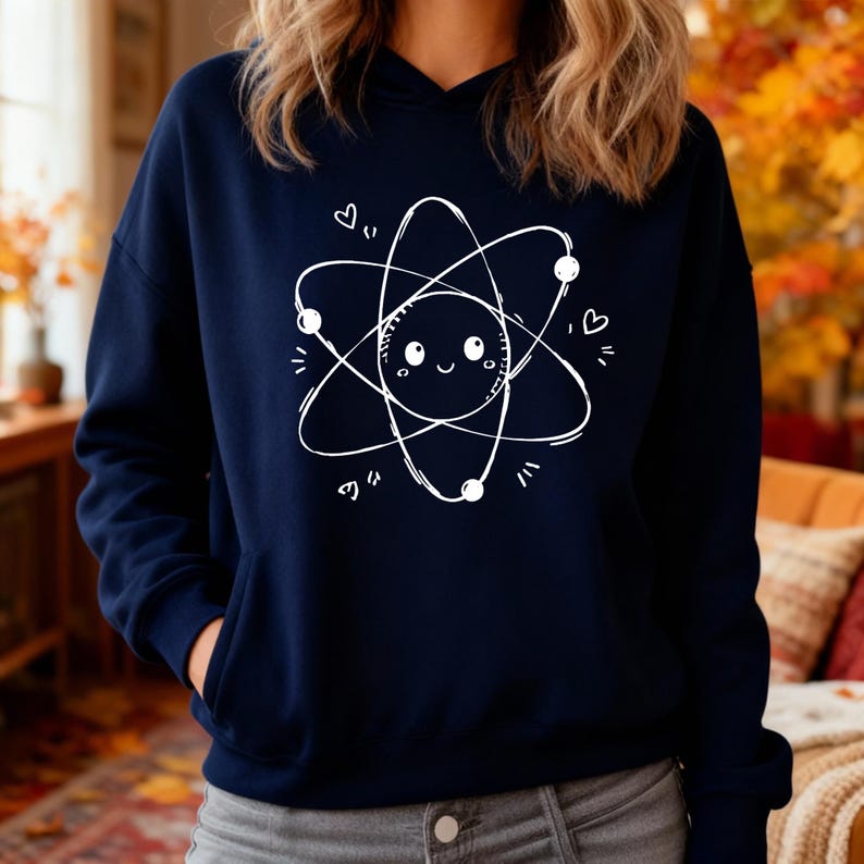 May include: A navy blue hooded sweatshirt with a white atom graphic. The atom design has a smiling face at the centre, with orbiting circles and dots, and small heart details. The sweatshirt has a front pocket and a hood.