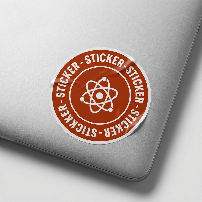 May include: A circular sticker with a white border and a rust-coloured centre. The centre features a white atom symbol. The words "STICKER" are printed in white around the edge of the sticker. The sticker is on a grey laptop.