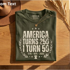 May include: An olive green t-shirt with the text "AMERICA TURNS 250 I TURN 50" in white, along with the years 1776-1976-2026. The shirt has a crew neck and is made in the USA. A gold necklace and a brown hat are also visible.
