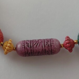 May include: Close-up of a colourful beaded necklace. The necklace features a central, cylindrical, carved purple bead, flanked by smaller, textured beads in shades of red, yellow, and green. Gold-coloured spacers separate the beads.