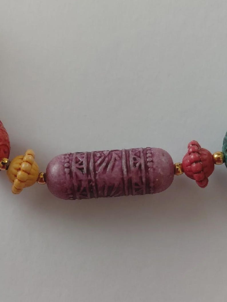 May include: Close-up of a colourful beaded necklace. The necklace features a central, cylindrical, carved purple bead, flanked by smaller, textured beads in shades of red, yellow, and green. Gold-coloured spacers separate the beads.