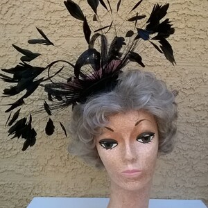 May include: A black feather hat with a pink base. The hat is on a mannequin head with gray hair.