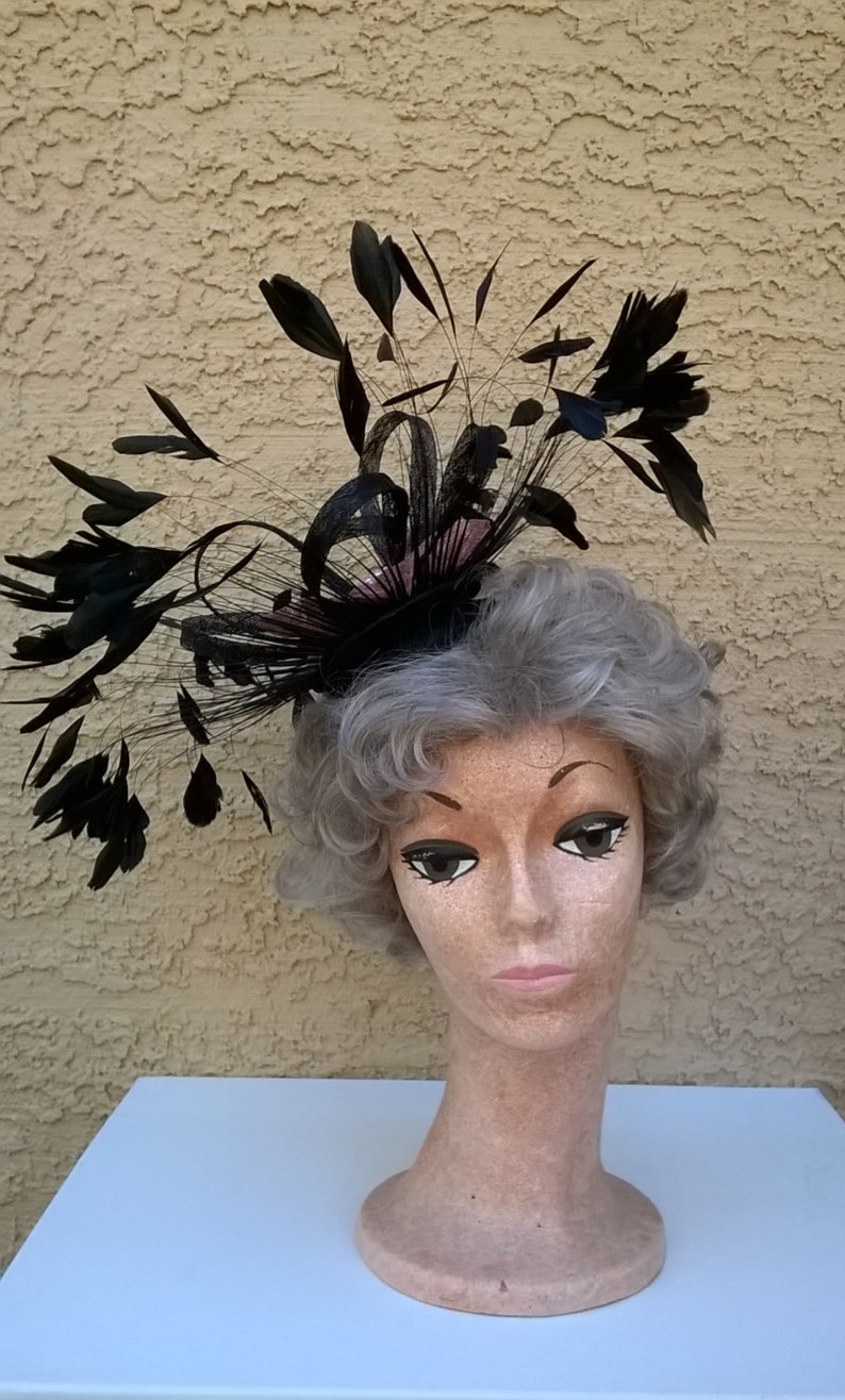 May include: A black feather hat with a pink base. The hat is on a mannequin head with gray hair.